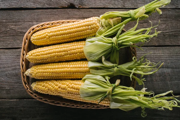 Draufsicht auf frische rohe Maiskolben im Korb auf hölzerner Tischplatte — Stockfoto