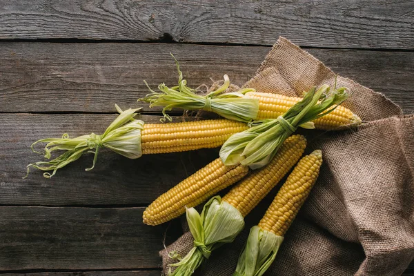 Draufsicht auf frische rohe Maiskolben auf Sacktuch auf hölzerner Tischplatte — Stockfoto