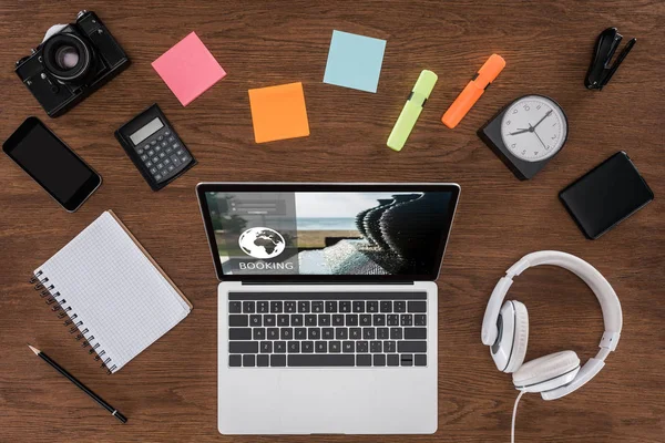 Draufsicht auf Holztisch mit leerem Lehrbuch, Smartphone, Fotokamera und Laptop mit Buchung auf dem Bildschirm — Stockfoto