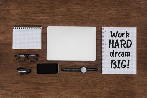 Vista superior del lugar de trabajo con ordenador portátil arreglado, reloj de pulsera, teléfono inteligente y libro de texto con letras trabajo duro sueño grande en la mesa de madera - foto de stock