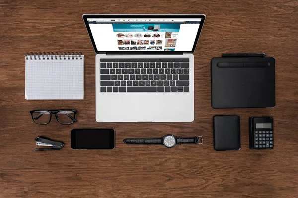 Vista superior del lugar de trabajo con libro de texto vacío arreglado, teléfono inteligente, reloj de pulsera y computadora portátil con Amazon en la pantalla - foto de stock