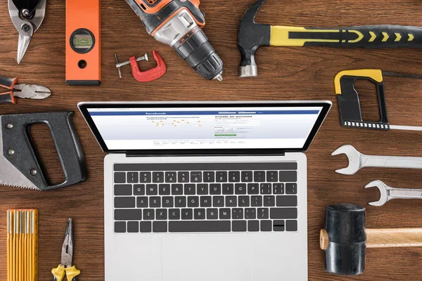 Ansicht des Arbeitsplatzes mit Laptop und Facebook auf dem Bildschirm, umgeben von verschiedenen Werkzeugen auf einem Holztisch — Stockfoto