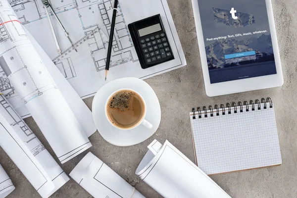 Вид сверху на рабочее место архитектора с чашкой кофе, чертежами, калькулятором и цифровым планшетом с tumblr на экране — стоковое фото