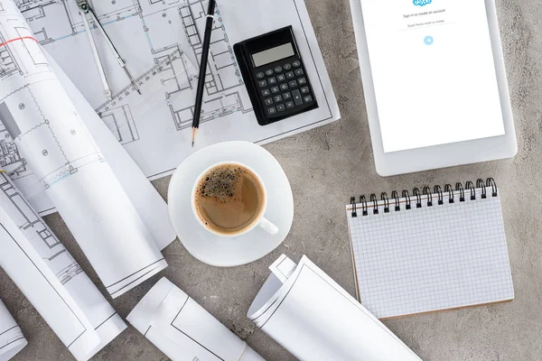 Vista superior del lugar de trabajo del arquitecto con taza de café, planos, calculadora y tableta digital con skype en la pantalla - foto de stock