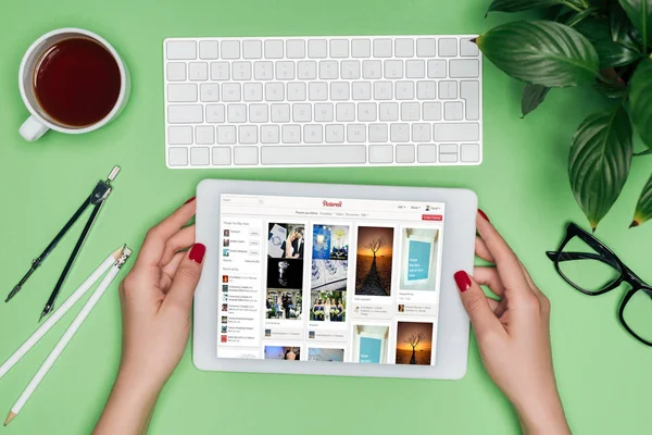 Cropped image of female architect holding digital tablet with pinterest on screen at table with divider, coffee and potted plant — Stock Photo