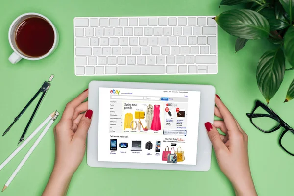 Cropped image of female architect holding digital tablet with ebay on screen at table with divider, coffee and potted plant — Stock Photo