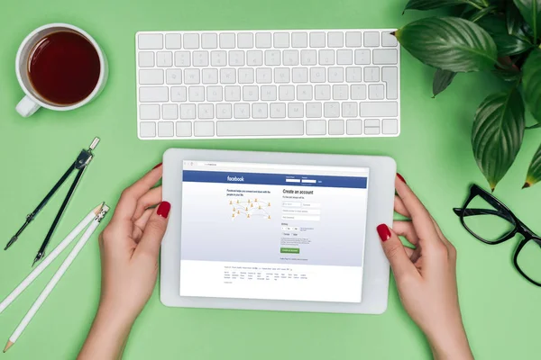 Cropped image of female architect holding digital tablet with facebook on screen at table with divider, coffee and potted plant — Stock Photo