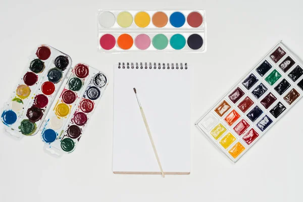 Top view of empty textbook and paintbrush surrounded by colorful paints on white table — Stock Photo