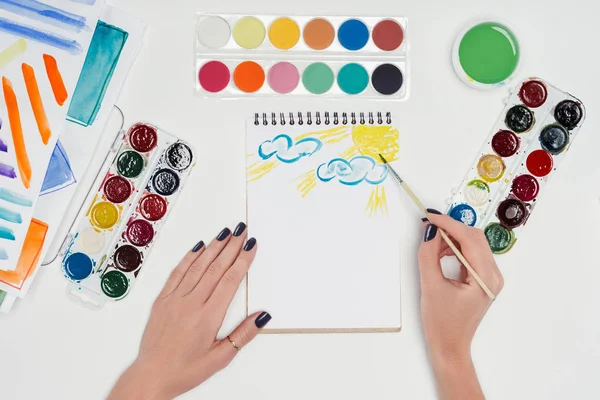 Partial view of female painter drawing by paintbrush in textbook at white table with colorful paints — Stock Photo