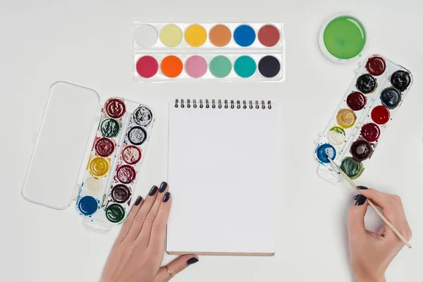 Zugeschnittenes Bild einer Künstlerin, die mit dem Pinsel in einem leeren Lehrbuch an einem weißen Tisch mit bunten Farben zeichnet — Stockfoto