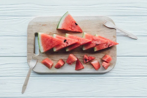 Flat lay with arranged watermelon pieces on cutting board and plastic forks on wooden tabletop — Stock Photo