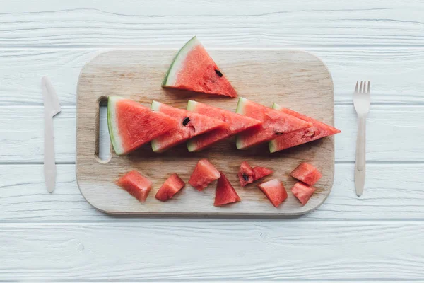 Flat lay with arranged watermelon pieces on cutting board and plastic cutlery on wooden tabletop — Stock Photo