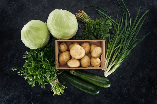 Top view of potatoes in box with arranged green vegetables on black marble surface — Stock Photo
