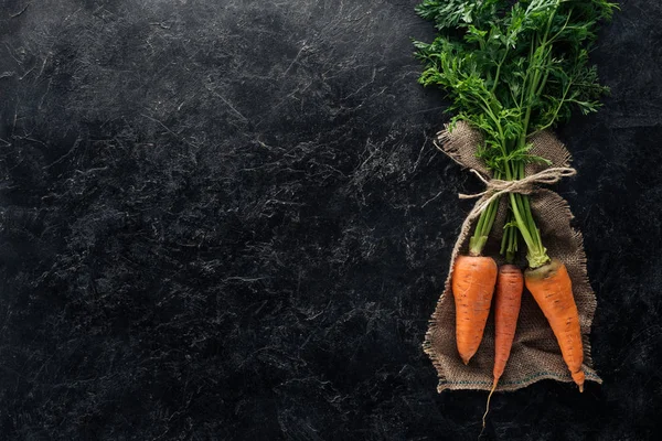 Vista superior de zanahorias frescas maduras atadas con cuerda sobre tela de saco sobre superficie de mármol negro - foto de stock