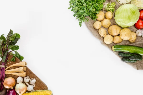 Vista superior de verduras dispuestas crudas en tabla de cortar de madera y saco aislado en blanco - foto de stock