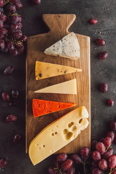 Vista superior del queso surtido en tabla de cortar de madera con uva en la superficie oscura - foto de stock