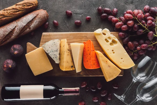 Acostado plano con baguettes, queso surtido, botella de vino y frutas en la mesa oscura - foto de stock