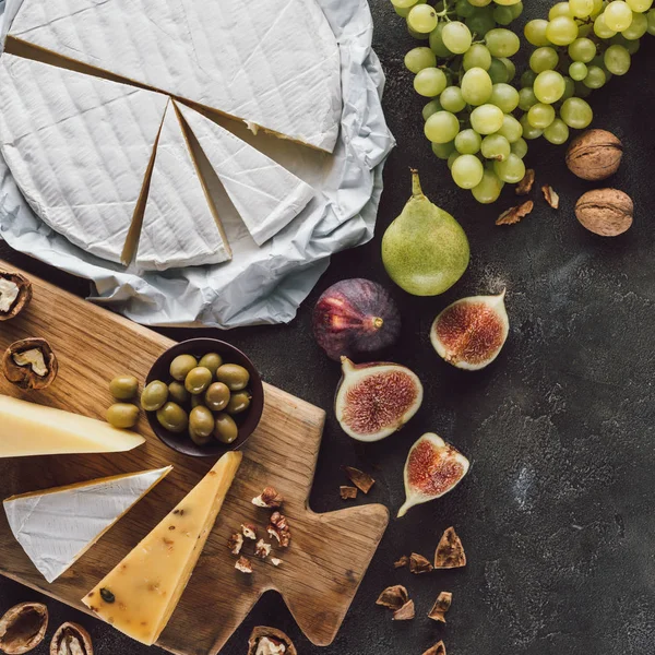 Вид сверху на разнообразный сыр, оливки в миске и фрукты на темной столешнице — стоковое фото