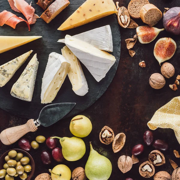 Flat lay with assorted cheese, olives in bowl and fruits on dark surface — Stock Photo