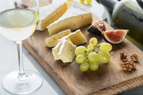 Vista de cerca de los trozos de queso camembert, uva e higo en la tabla de cortar con un vaso de vino cerca en la mesa blanca - foto de stock