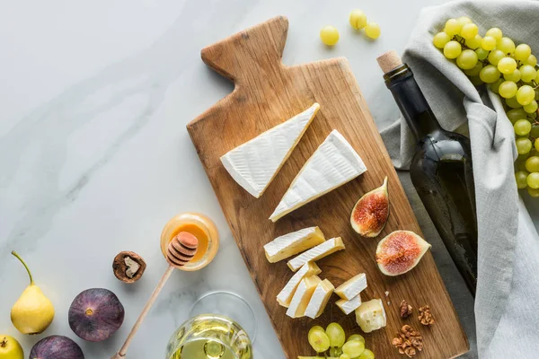Flache Lage mit Essenszusammensetzung aus Käse auf Schneidebrett, Honig, Wein und Früchten auf weißer Marmoroberfläche — Stockfoto