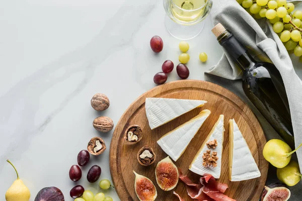 Flat lay com vinho, queijo, jamon e várias frutas dispostas em tábua de madeira em mesa de mármore branco — Fotografia de Stock