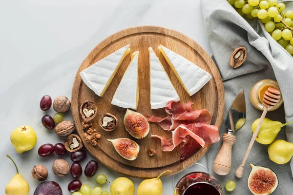 Flat lay with cheese, jamon, figs, hazelnuts and grape on wooden board on white marble tabletop — Stock Photo