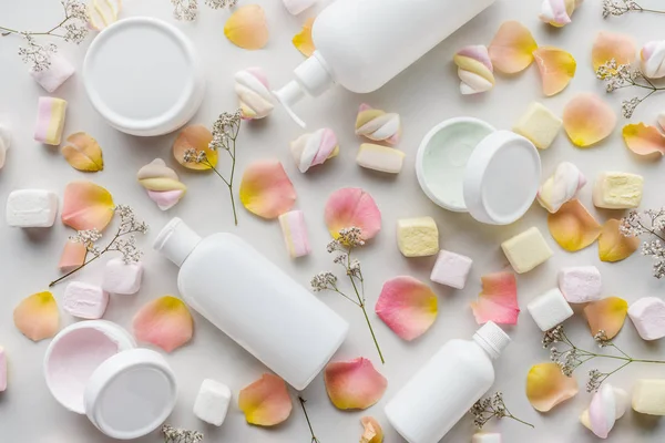 Top view of bottles of cream, rose petals and marshmallows on white tabletop, beauty concept — Stock Photo