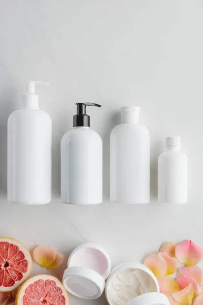 Vista superior de botellas de crema, pedazos de pomelo y pétalos de rosa en la superficie blanca, concepto de belleza - foto de stock