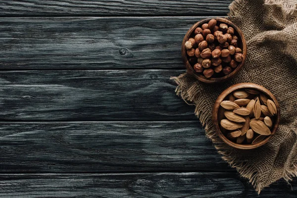 Almonds and hazelnuts in wooden bowls with sackcloth on dark wooden surface — Stock Photo