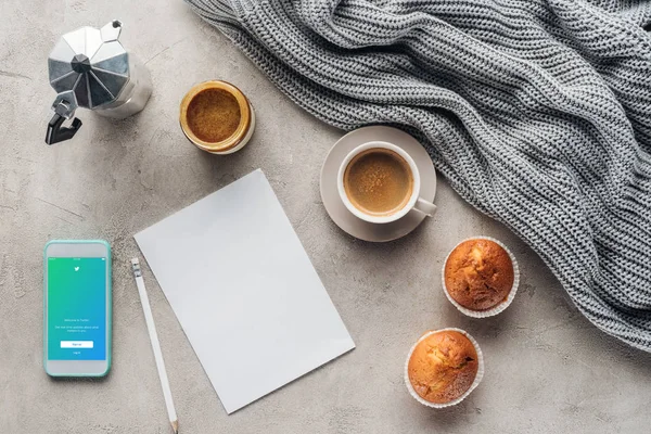 Top view of cup of coffee with muffins, blank paper and smartphone with twitter app on screen on concrete surface with knitted wool drapery — Stock Photo
