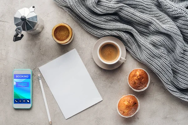 Vista superior de la taza de café con magdalenas, papel en blanco y teléfono inteligente con aplicación de reserva en la pantalla en la superficie de hormigón con cortinas de lana de punto - foto de stock