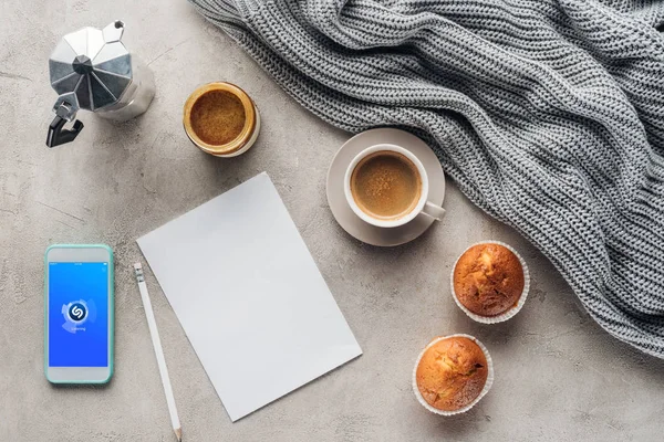 Top view of cup of coffee with muffins, blank paper and smartphone with shazam app on screen on concrete surface with knitted wool drapery — Stock Photo