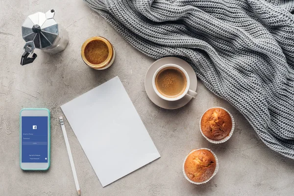 Top view of cup of coffee with muffins, blank paper and smartphone with facebook app on screen on concrete surface with knitted wool drapery — Stock Photo