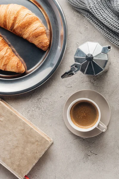 Top view of cup and moka pot of coffee with book and croissants on concrete surface — Stock Photo