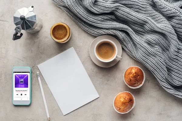 Top view of cup of coffee with muffins, blank paper and smartphone with music player app on screen on concrete surface with knitted wool drapery — Stock Photo