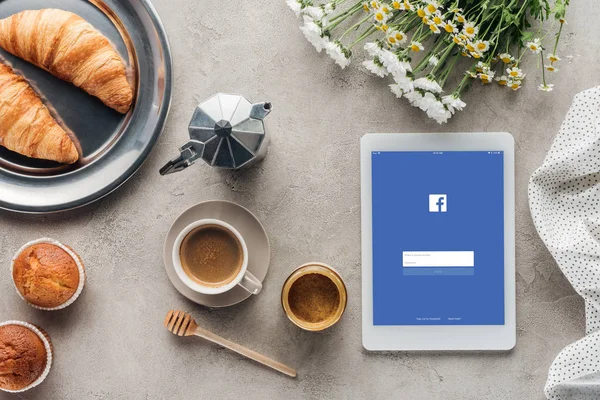 Верхний вид кофе с выпечкой и планшетом с приложением facebook на экране на бетонной поверхности — стоковое фото