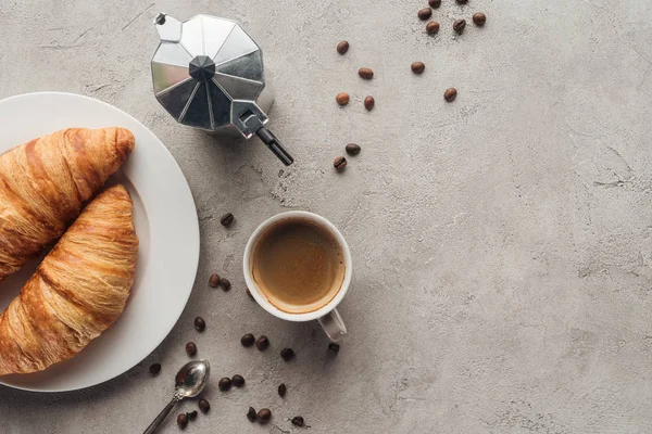 Draufsicht auf Tasse Kaffee mit Croissants und Mokka-Kanne auf Betonoberfläche mit verschütteten Kaffeebohnen — Stockfoto