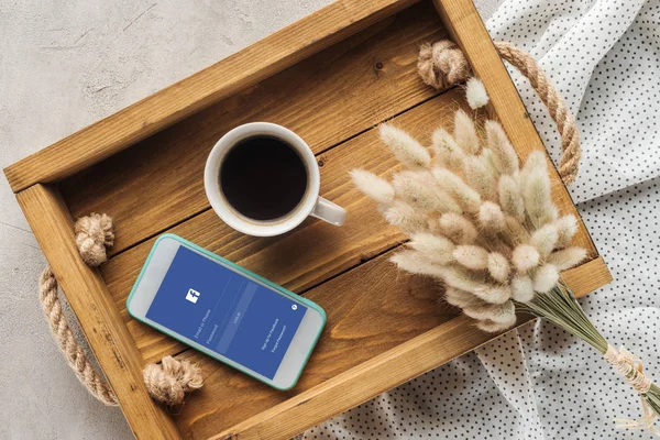 Draufsicht auf Tasse Kaffee und Smartphone mit Facebook-App auf dem Bildschirm auf Tablett mit Lagurus ovatus Bouquet auf Betonoberfläche — Stockfoto