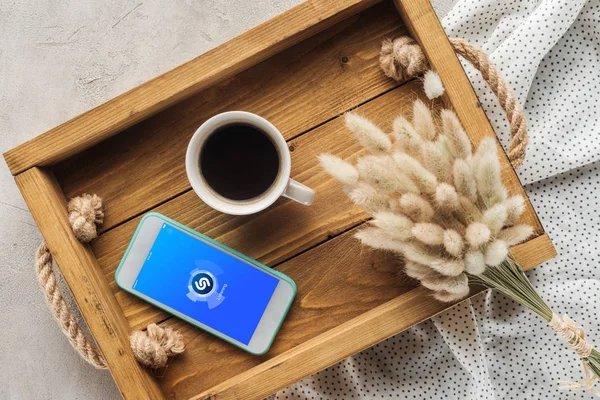 Draufsicht auf Kaffeetasse und Smartphone mit Shazam-App auf dem Bildschirm auf Tablett mit Lagurus ovatus Bouquet auf Betonoberfläche — Stockfoto