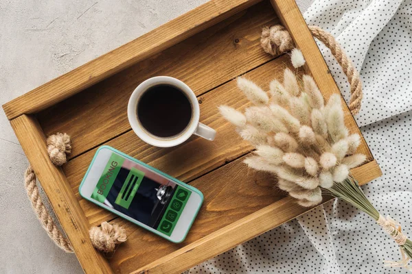 Top view of cup of coffee and smartphone with booking website on screen on tray with lagurus ovatus bouquet on concrete surface — Stock Photo