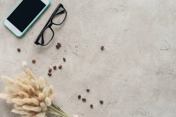 Vista dall'alto dello smartphone con schermo bianco con occhiali, chicchi di caffè rovesciato e bouquet lagurus ovatus sulla superficie in cemento — Foto stock
