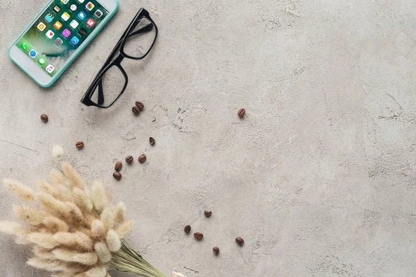Вид сверху на смартфон с ios домашним экраном с очками, пролитыми кофейными зёрнами и букетом Lagurus ovatus на бетонную поверхность — стоковое фото