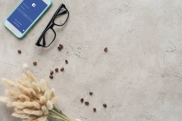 Вид сверху на смартфон с приложением facebook на экране с очками, пролитыми кофейными зёрнами и букетом лагурус оватус на бетонной поверхности — стоковое фото