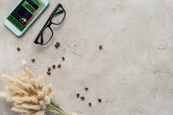 Vue du haut du smartphone avec application de réservation à l'écran avec lunettes, grains de café renversés et bouquet lagurus ovatus sur la surface du béton — Photo de stock