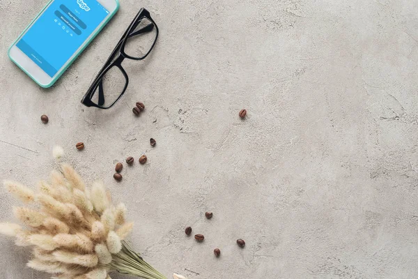 Top view of smartphone with skype app on screen with eyeglasses, spilled coffee beans and lagurus ovatus bouquet on concrete surface — Stock Photo