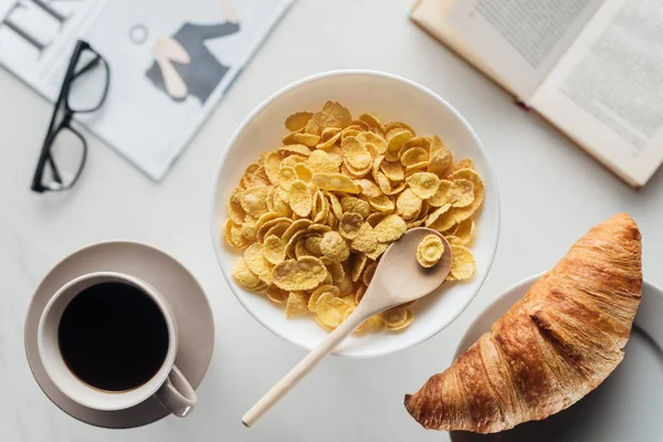 Top view of bowl of dry cereal breakfast with cup of coffee and croissant on white surface with newspaper and book — Stock Photo