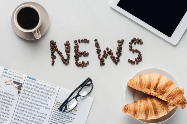 Vista superior del café con letras de noticias hechas de granos de café, croissants y periódico con la tableta en mármol blanco - foto de stock