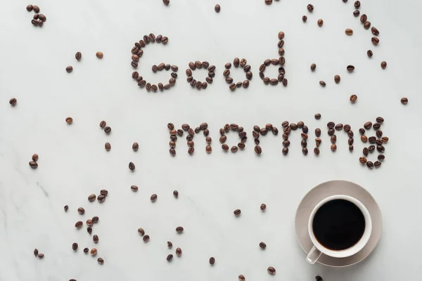 Draufsicht auf Tasse leckeren Kaffee und Guten Morgen Schriftzug aus Kaffeebohnen auf weißem Marmor — Stockfoto