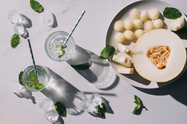 Top view of sweet melon balls, half of ripe melon and refreshing beverage with mint and drinking straws in glasses on white — Stock Photo